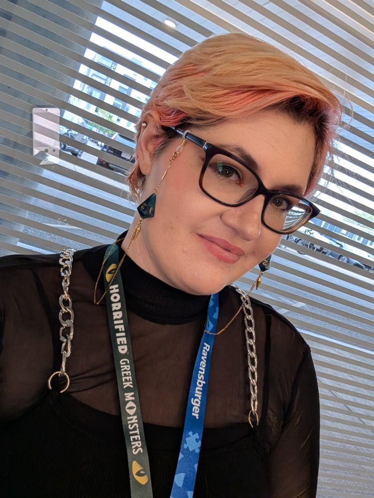 A feminine presenting person with blonde/pink hair and glasses with dice chains on it sitting in front of a window.