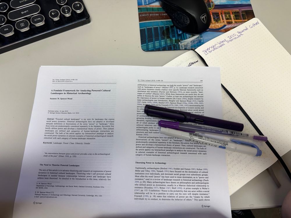 A printed article titled “a feminist framework for analyzing powered cultural landscapes in Historical Archaeology” with a gelly roll pen, a lamy safari pen, and a zebra mildliner on top of it on a background of a computer desk. A keyboard is visible in the background.