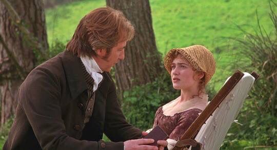 a man with long hair and a brown coat with a small book in his hand sits by a woman in a brown hat.