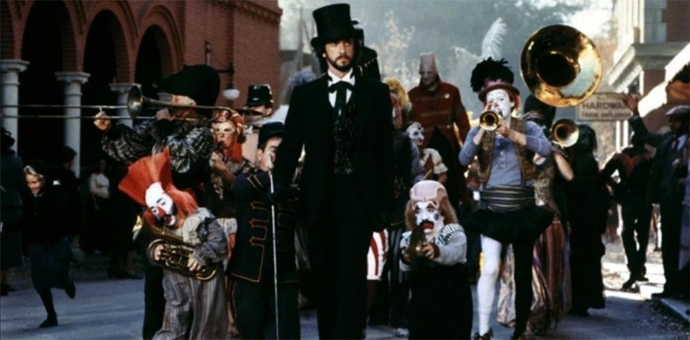 a man in a dark suit, top hat and cane walks down a village street before a group of clowns.  There's a tuba at the back.