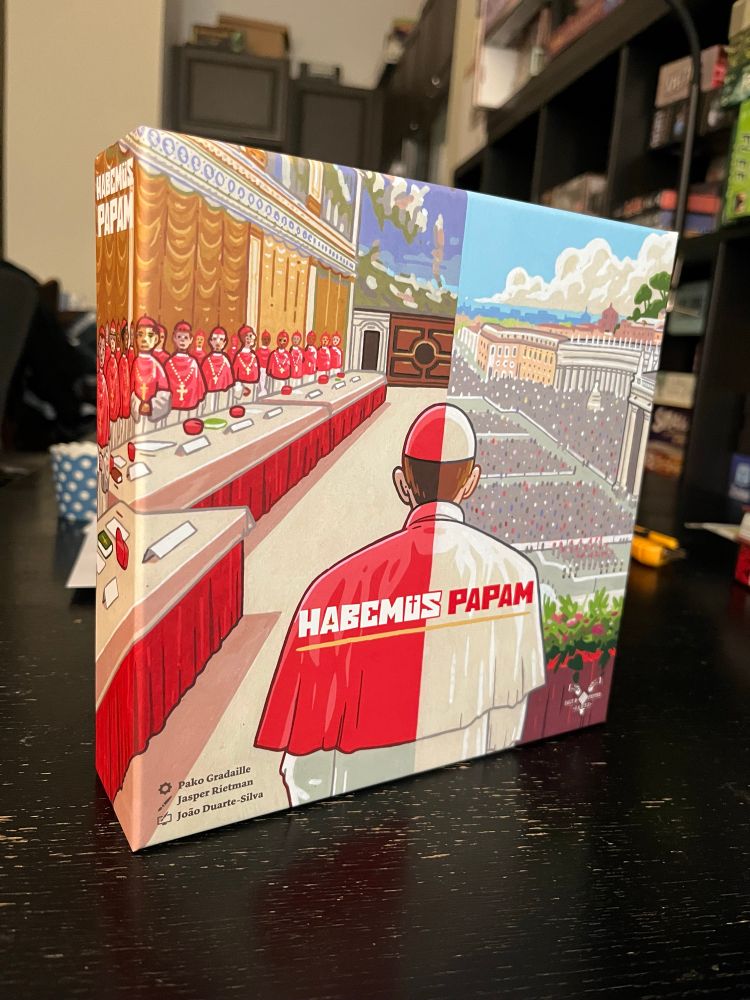 Box cover for Habemus Papem tabletop game, showing a split-screen cardinal looking at a council on the left and St Peter’s Square on the right. 