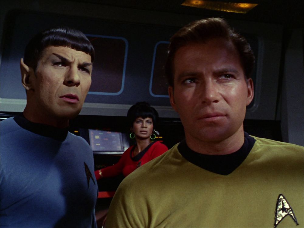 A still from the episode "A Balance of Terror"; Kirk and Spock sand on the foreground, Spock to the left and Kirk to the right. A shadow covers half of Kirk's face as both he and Spock look towards the viewscreen, off-camera. Framed in-between the two men on the background, Uhura, on her bridge station, also looks towards the viewscreen. All display faces of professionally contained tension.