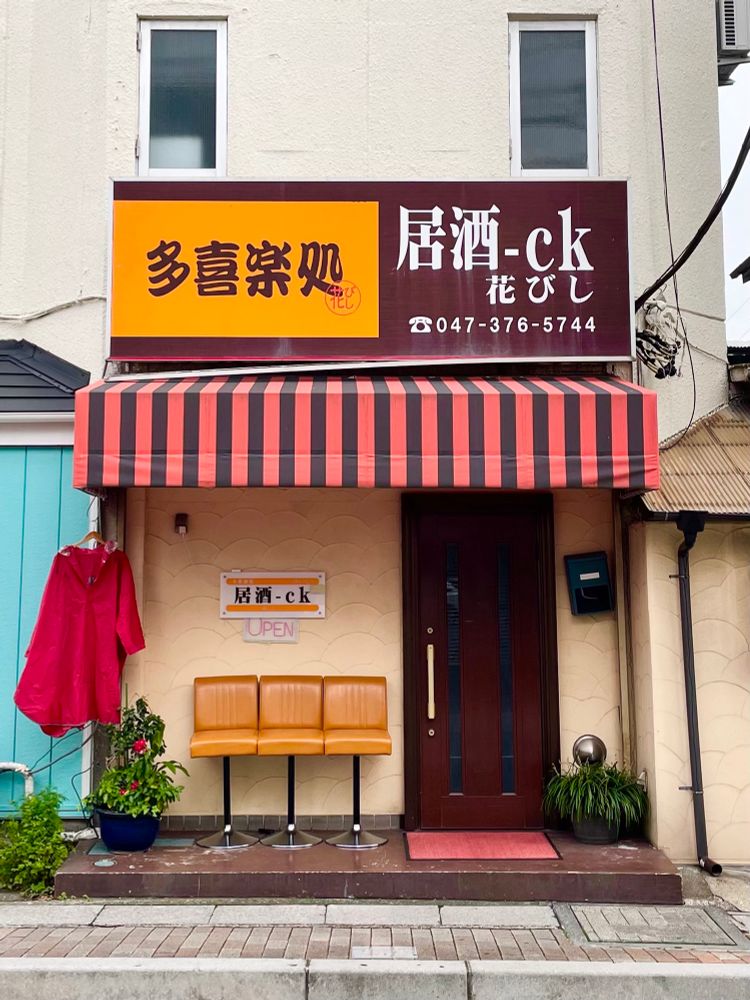 An izakaya with striped awning, three Shōwa era bar stools  lined up outside and a red cagoule hanging to dry