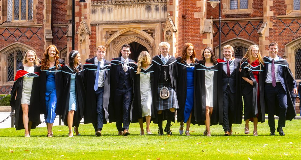 Image shows 12 classmates from the School of Medicine, Dentistry and Biomedical Sciences who graduated today with a degree in Medicine. They are walking arm in arm across the lawn in front of the Lanyon Building.