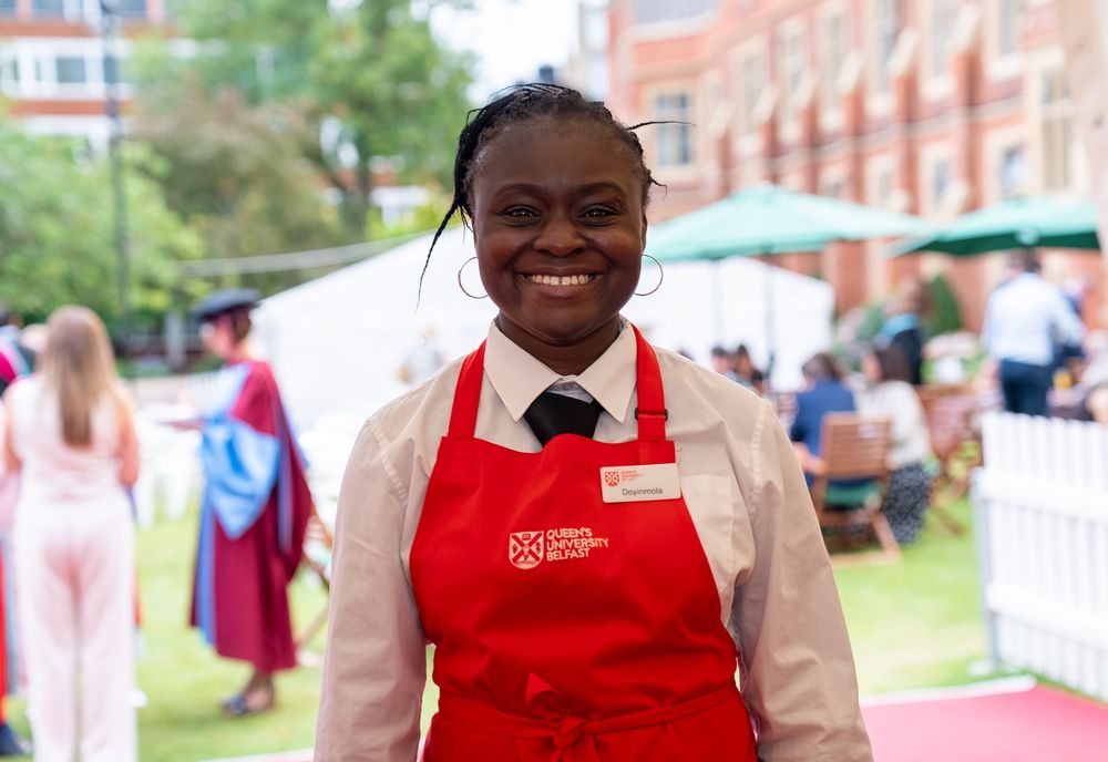 Image shows a member of waiting staff in the Graduation marquee. In the background there are tables and umbrellas and people socialising.