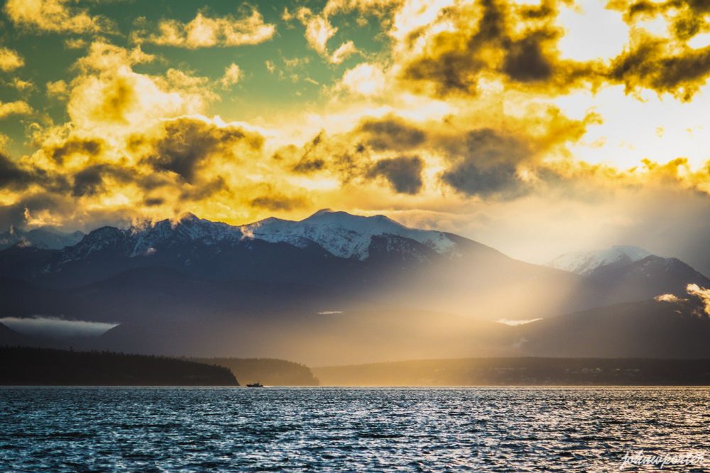 A small boat floats on Puget Sound at sunset, with dramatic dark clouds above and golden sunlight breaking through. The Olympic Mountains form a distant silhouette.