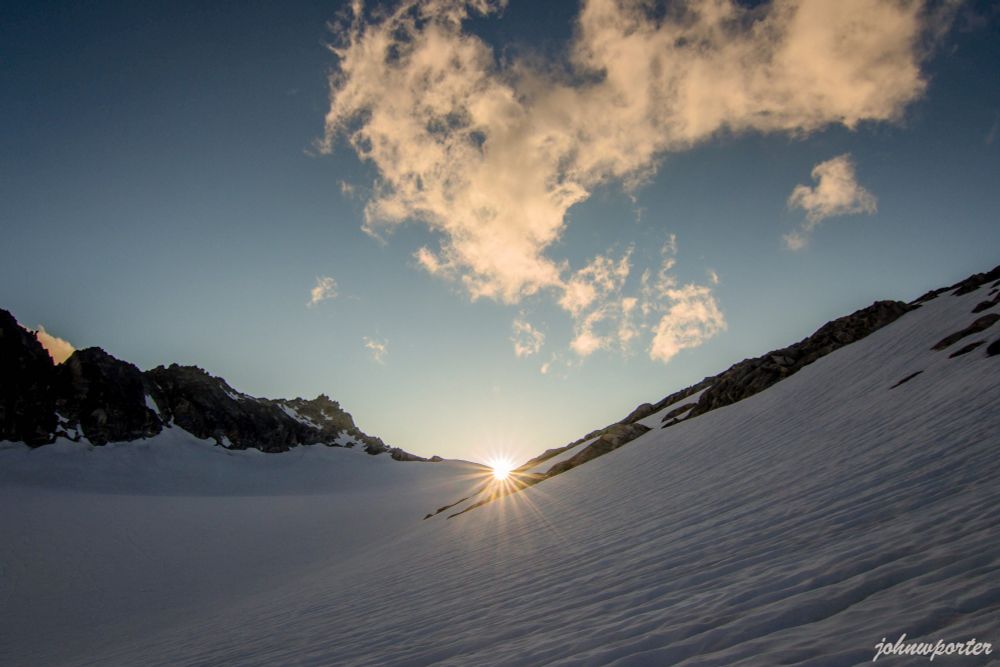 Sunset casts light from the west on the ridgeline of Primus Peak, visible from North Klawatti Glacier Camp. The hues illuminate wispy, hovering clouds under a darkening sky in the North Cascades National Park.