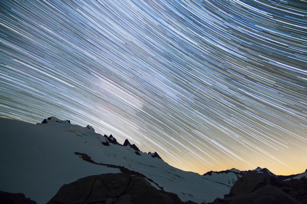 Star trails streak across the night sky, creating luminous arcs above the snow-capped Mount Challenger of Northern Picket Range, perching above the Challenger Glacier.