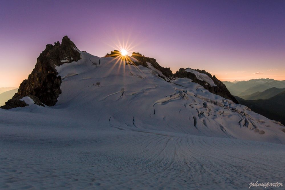 Sunburst crests the jagged ridgeline of Dorado Needle, visible from Tepeh Col in the east. The light casts a warm glow on McAllister Glacier and the surrounding ice in the North Cascades National Park. 