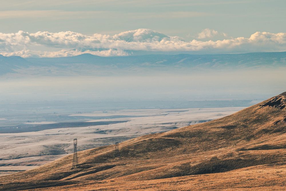 A distant view from Rattlesnake Hills in Eastern Washington, across a frosty, rolling landscape toward a cloud-covered Mount Rainier on the horizon. Layers of hills fade into the background under a pale blue sky, with golden-brown slopes in the foreground and faint power lines cutting across the open terrain.