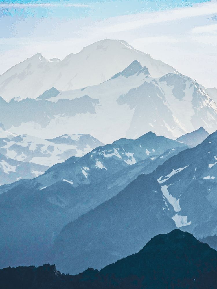 A long-distance, hazy shot of the layered Cascade Mountains taken from Hozomeen Mountain South Peak near the Canadian border by Ross Lake. Mount Shuksan rises in the mid-ground and the larger Mount Baker stands prominently in the distance against a light blue sky.