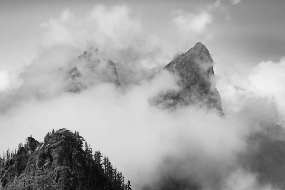 A dramatic black and white picture featuring the two angular summits of Baring Mountain emerging from a thick blanket of clouds. In the foreground is a ridge covered in burned trees, captured from Grotto Mountain across the South Fork Skykomish River Valley.
