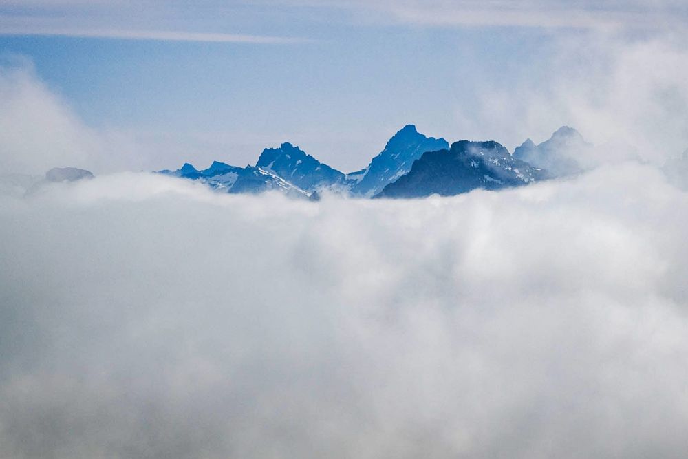 Emerging from a soft, undulating layer of clouds, the sharp silhouettes of Overcoat Peak and Chimney Rock in the Alpine Lakes Wilderness stand tall and imposing against the sky.