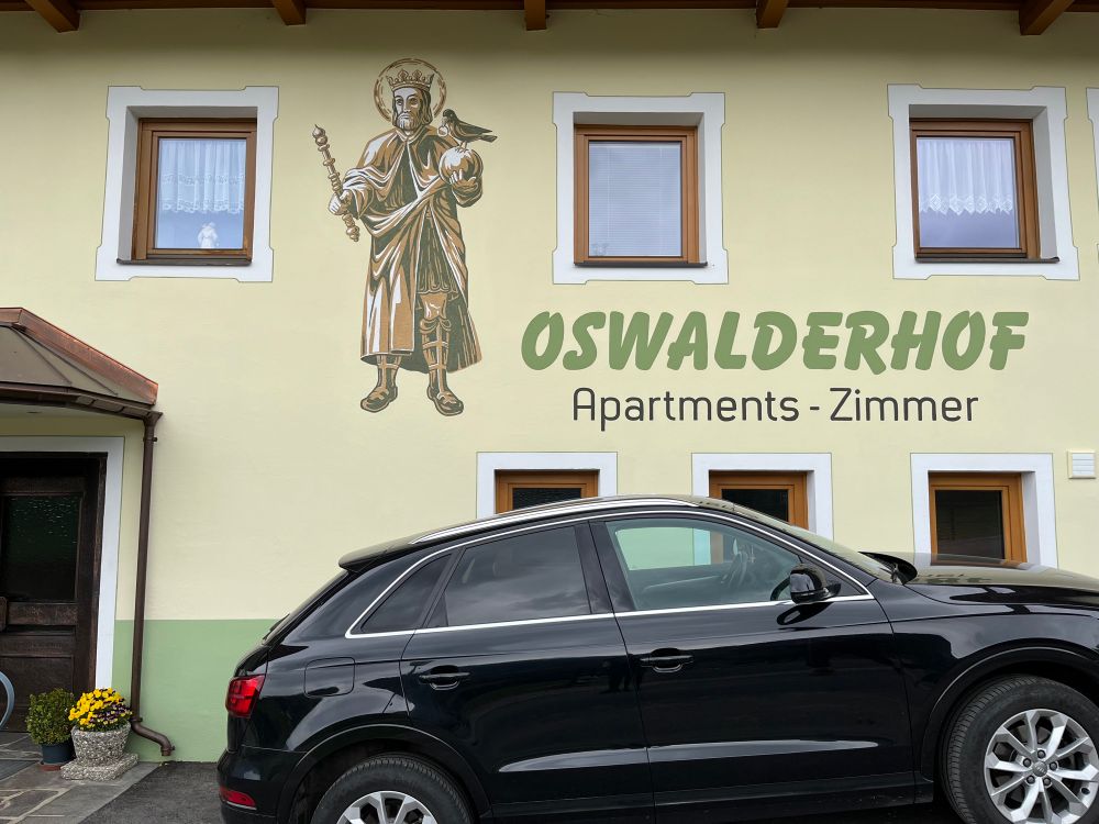 Holiday apartments called the Oswalderhof with a painting of a saint king holding a sceptre and an orb with a Raven holding a ring in its beak perched on the orb.