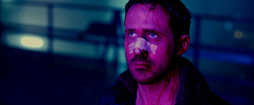 A shot of K from Blade Runner 2049, played by Ryan Gosling. He is bathed in soft pink and purple light from the front, and stares up into the middle distances he carries a hollow look in his blue eyes, is covered in blood, and has a white bandage on his nose.