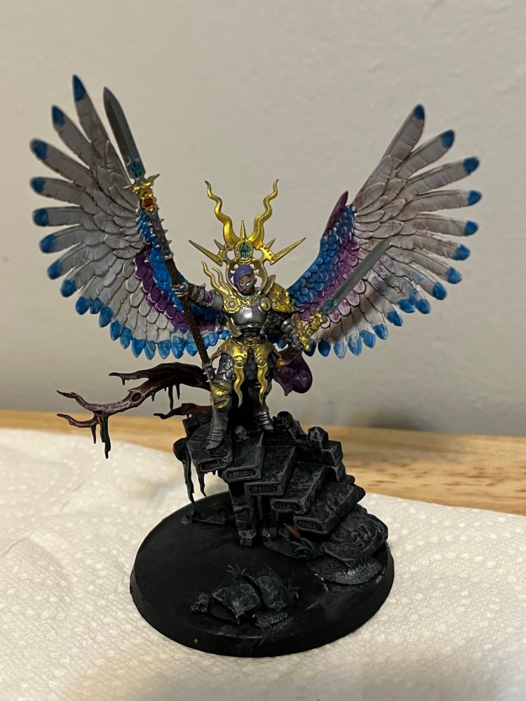 Yndrasta, the Celestial Spear from Warhammer: Age of Sigmar. 

Yndrasta is an angel with silver base armor, golden pauldrons, the pauldron on her left shoulder is a sculpted bearded man. Gold armor pieces rest outside her chain mail skirt, she has blue, white, and purple wings. The wing tips are a blue, and the top of her wings are blue fading to purple with large swaths of white before the tips. They are outstretched, as if she is taking flight or posing to intimidate with her wings.

Yndrasta stands on the top of a ruined dark grey spiral staircase, a skull resting to the right of her left leg. A dark brown tree juts out behind the ruined stair case, and vines drape and grow down multiple stairs. 

She poses with two weapons outstretched, in her right: a spear larger than her body, silvered blade, with a glowing turquoise rune, gold hilt with an inset ruby that connects to a long leathery spear shaft.

In her left hand: a long sword, with a similar turquoise rune inset, ruby inlaid in the hilt, and leather wrapped handle, capped with a gold pommel.

Yndrasta’s face is dark skinned, her eyes glow white with blue lighting streaks bleeding onto her face. Her hair is in a half shaved style, with purple and blue hair transitioning to a very small black root that drapes chin length over the right side of her face.

Behind Yndrasta set in the armor is a gold crown wrapped around her armor shooting out spikes like lightning or sunbursts , with an inlaid turquoise rune.