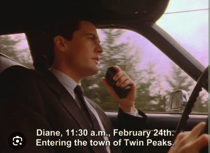 Still of Agent Dale Cooper driving in his car while holding a voice recorder in one hand, subtitled, 'Diane, 11:30 a.m., February 24th. Entering the town of Twin Peaks.'