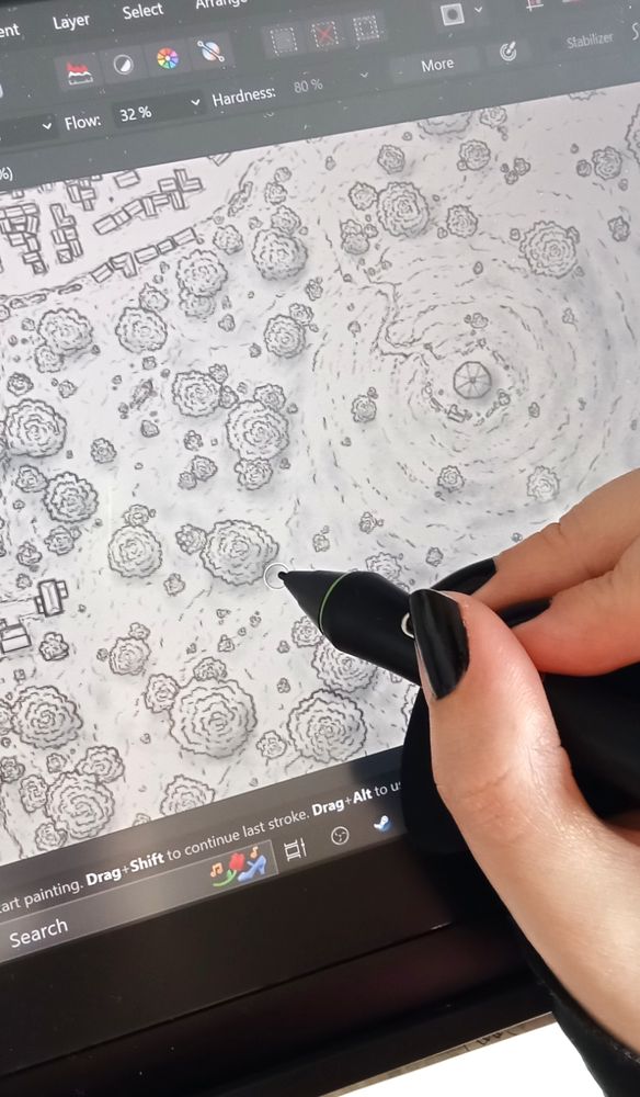 A close up view of a hand holding a wacom stylus, working on a black and white drawing of a map showing a section with lots of trees and some buildings in the top corner, and a hill to the right with a tower and some broken walls around it.