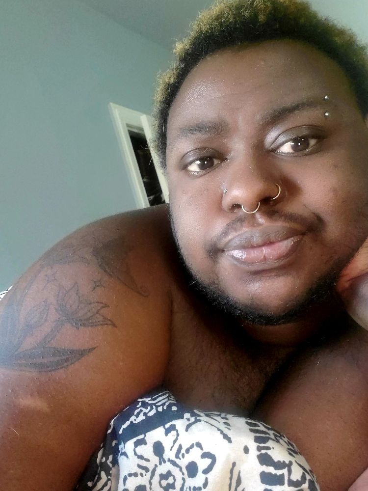 A fat dark skinned Black person with faded yellow hair and beard is looking into the camera. He has a nose stud in one nostril, a hoop in the other and a septum. His forearm tattoo is of a flower.
