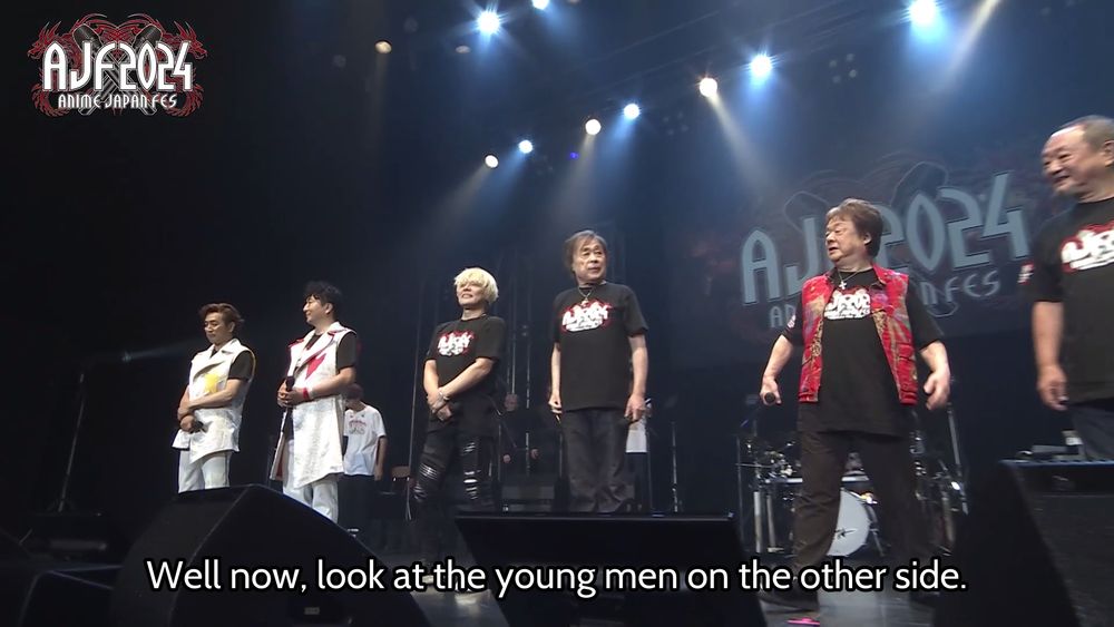 A shot of a bunch of singers lined up on stage, with a subtitle of the MC saying that all of the young singers are on the opposite side of the stage as him.