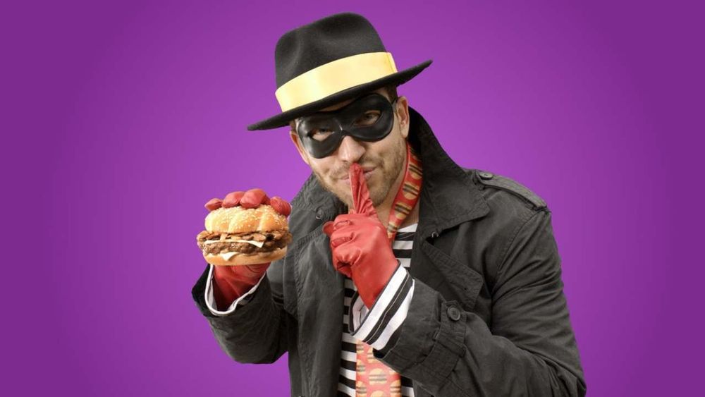 an image of the mcdonaldland character hamburglar, reimagined in 2015 as some kind of suburban dad, dressed as some kind of gender-swapped carmen sandiego, holding a double cheeseburger, all set against a purple background
