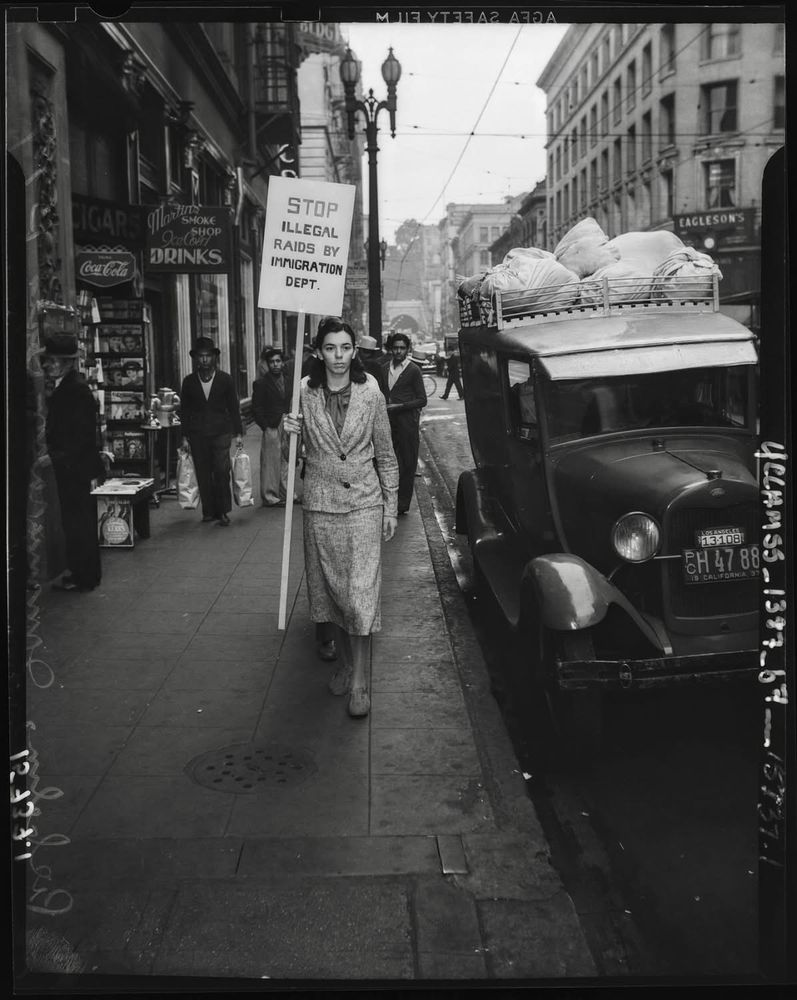 A black and white photo of a woman carrying a placard reading “stop illegal raids by immigration dept”