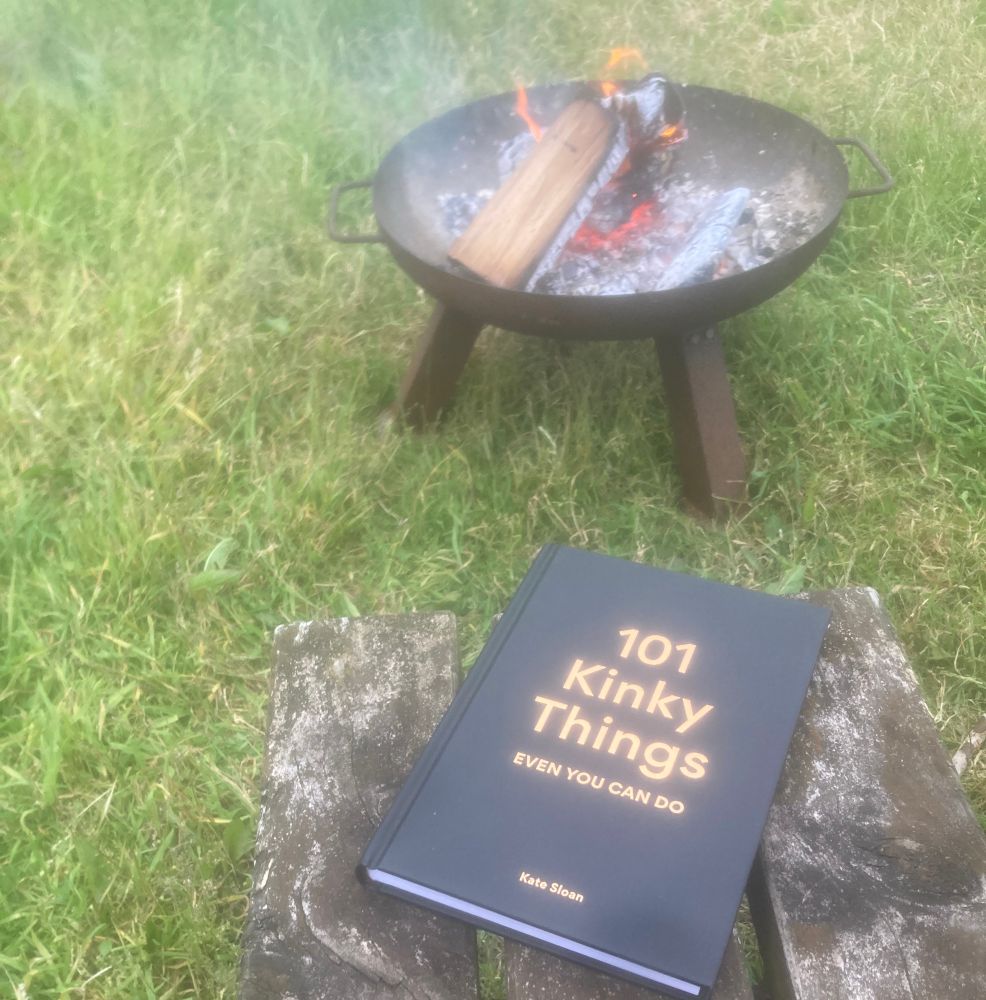 A book (101 kinky things even you can do by Kate Stern) on a bench next to an open fire in a raised iron fire pit in a grassy field.