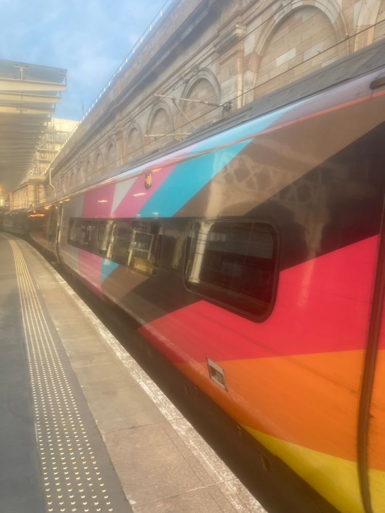 A pendolino train belonging to Avanti West Coast and sporting progress flag livery. The train is stopped at platform 9, Waverley Railway Station, Edinburgh, Scotland. It is a sunny morning and the train looks happy and excited for its journey (if you use your imagination).