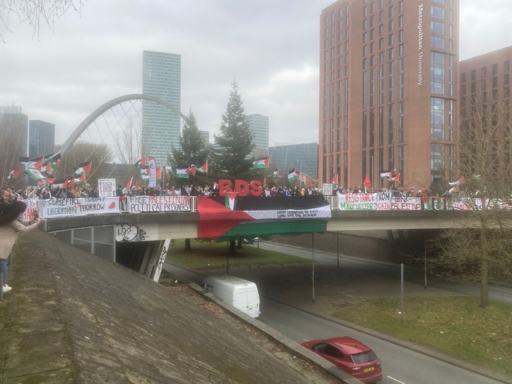 A banner drop on a bridge in Manchester. Visible are hundreds of people, Palestinian flags being waved, a giant Palestinian flag hanging from the bridge, also hanging from the bridge are banners reading: “ceasefire today, liberation tomorrow”; “free Palestinian political prisoners”; “resistance from Manchester to Gaza”; “honk for Palestine”; “stop Israel ethnic cleansing Gaza”.