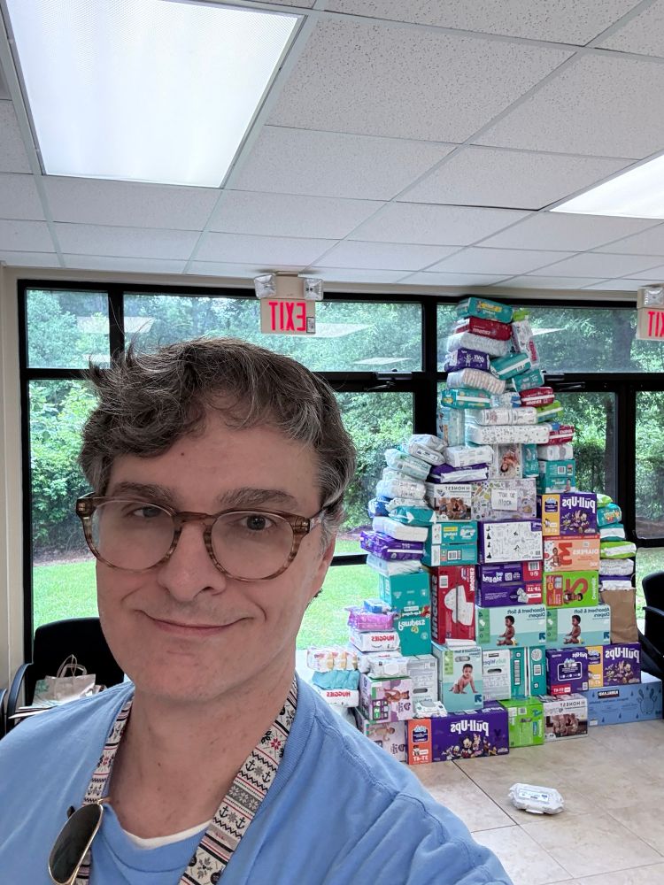 A smiling person wearing glasses and a blue shirt stands indoors in front of a large pyramid-shaped stack of donated diaper boxes, arranged like a Christmas tree. The room has large windows, a tiled floor, and a visible green lawn outside. Ceiling lights and EXIT signs hang above.