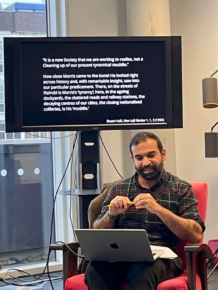 Photo of Nasser Mufti speaking while sitting on a chair in front of a screen with a quotation:
‘ It Is a new Society that we are working to realise, not a Cleaning up of our present tyrannical muddle.*
How close Morris came to the bone! He looked right across history and, with remarkable Insight, saw into our particular predicament. There, on the streets of Nairobi is Morris's tyranny: here, in the ageing dockyards, the cluttered roads and rallway stations, the decaying centres of our cites, the closing nationalised collieries, is his muddle.’
Stuart Hall, New Left Review 1. 1. 3 (1960)