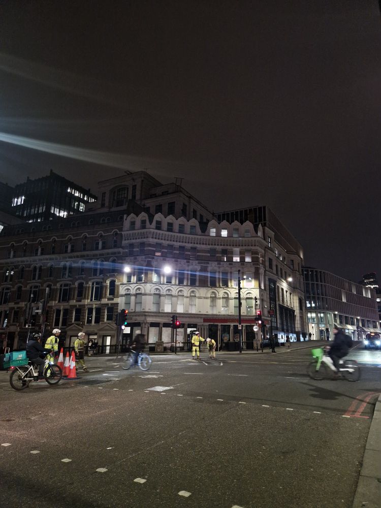 Workers in the middle of an intersection clearing something underground at the Blackfriars intersection with Farringdon. Two cyclists stream past in a blur.