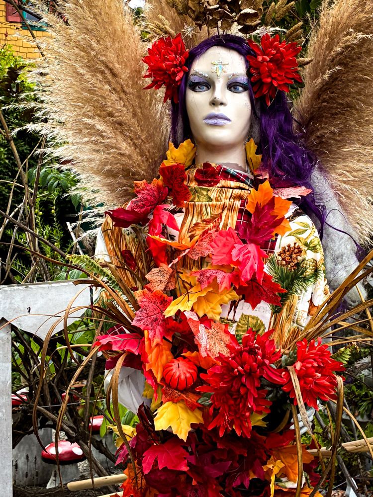 A scarecrow in the style of a woman with purple hair and flowers in her hair. She holds an arrangement of flowers. 