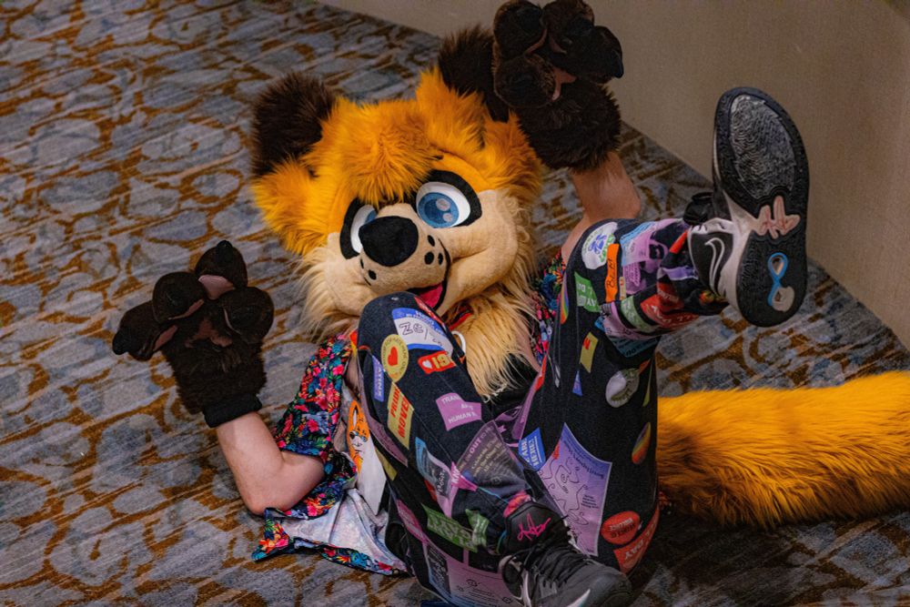 Person in fox fursuit, laying on ground at bottom of flight of stairs, legs and arms in air in an action pose, implying that they had just fallen down a flight of stairs. 