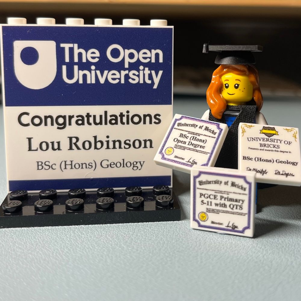 A close-up photo of a custom LEGO-style display celebrating a graduation. The display features a blue and white sign with “The Open University” logo and the text: “Congratulations Lou Robinson, BSc (Hons) Geology.” Next to the sign is a LEGO minifigure with long orange hair wearing a black graduation cap and gown. The minifigure is holding three small, tile-like certificates: one says “University of Bricks BSc (Hons) Open Degree,” another reads “University of Bricks BSc (Hons) Geology,” and the last one says “University of Bricks PGCE Primary 5-11 with QTS.” The background includes shelves with indistinct objects.