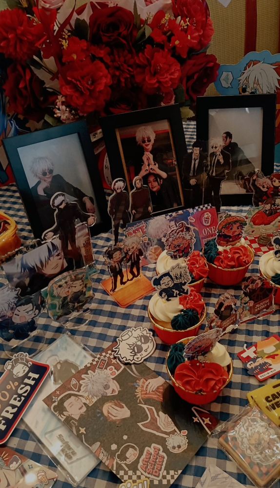 Decorated table with Gojo Satoru and Geto Suguru from jujutsu kaisen. The table is full of merchandises like prints, acrylic keychains and standees, stickers, cupcakes, cosplay photos and a bloombox full of flowers