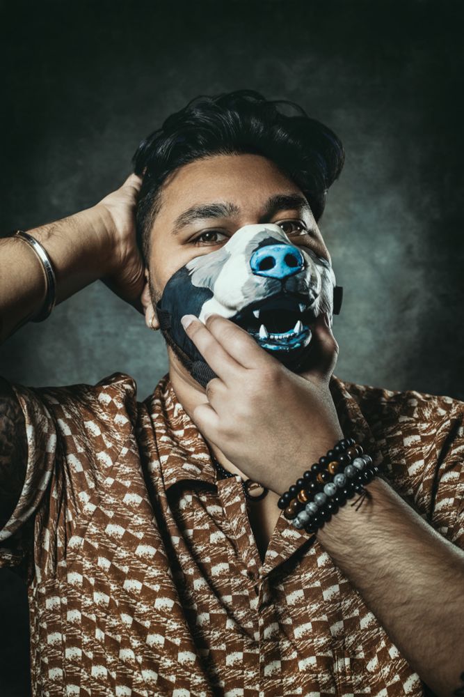 Valiant is a desi man wearing a print shirt, medium-length wavy hair and stubble. He is wearing a bear mask.