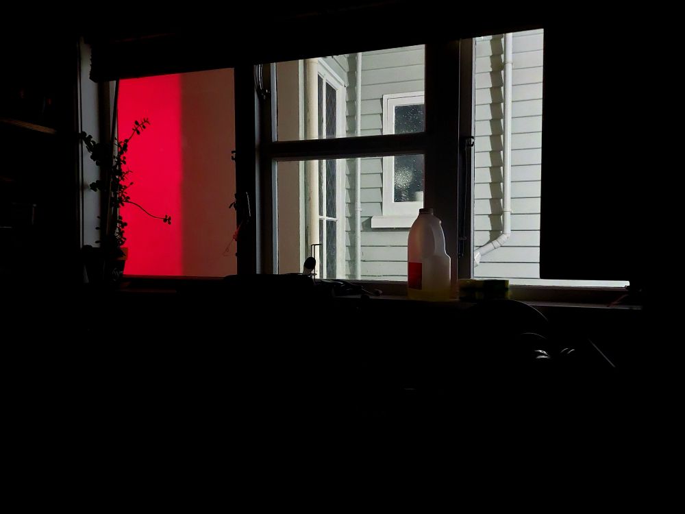 A view from a kitchen window looking out onto the wall of a neighbor's house, one third of which is lit blood red.