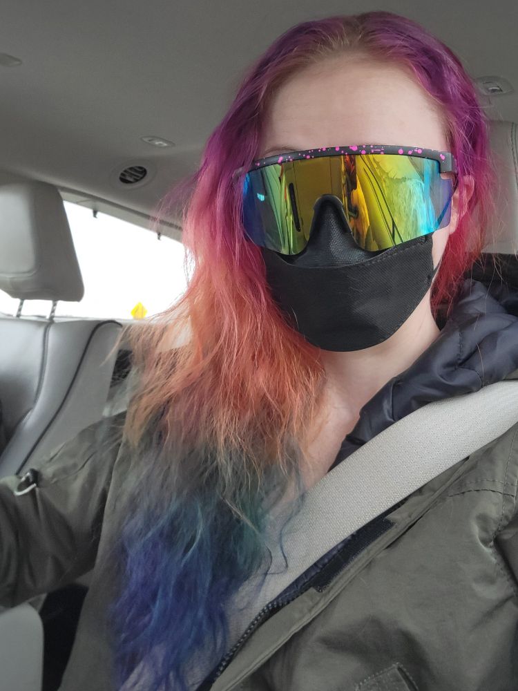 Lulu, with long rainbow hair and big reflective rainbow sunglasses and a black kn95 mask on in a car