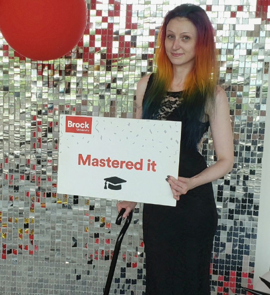 Lulu, pale woman with rainbow hair, wearing black dress and black cane with a sign that says "mastered it"
