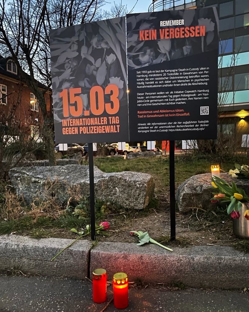 Das Foto zeigt einen Ausschnitt des Gedenkortes am 15.03. Zu sehen sind zwei Gedenkplakate, zu dessen Füßen Blumen liegen und zwei angezündete Grabkerzen stehen. Auf den Plakaten ist zu lesen: 15.03 internationaler Tag gegen Polizeigewalt. Sowie: Remember. Kein Vergessen. 
