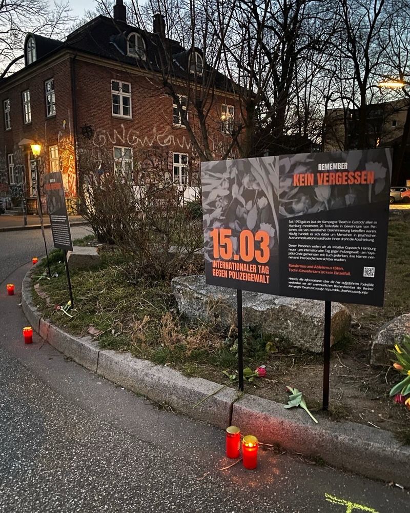 Das Foto zeigt einen Ausschnitt des Gedenkortes am 15.03. Zu sehen sind einige der Gedenkplakate, die kreisförmig entlang der Verkehrsinsel (Yaya Jabbi Circle) in der Erde stecken. Die Plakate sind an schwarzen Stöckern befestigt. Zu ihren Füßen liegen Blumen sowie entflammte rote Grablichter. 