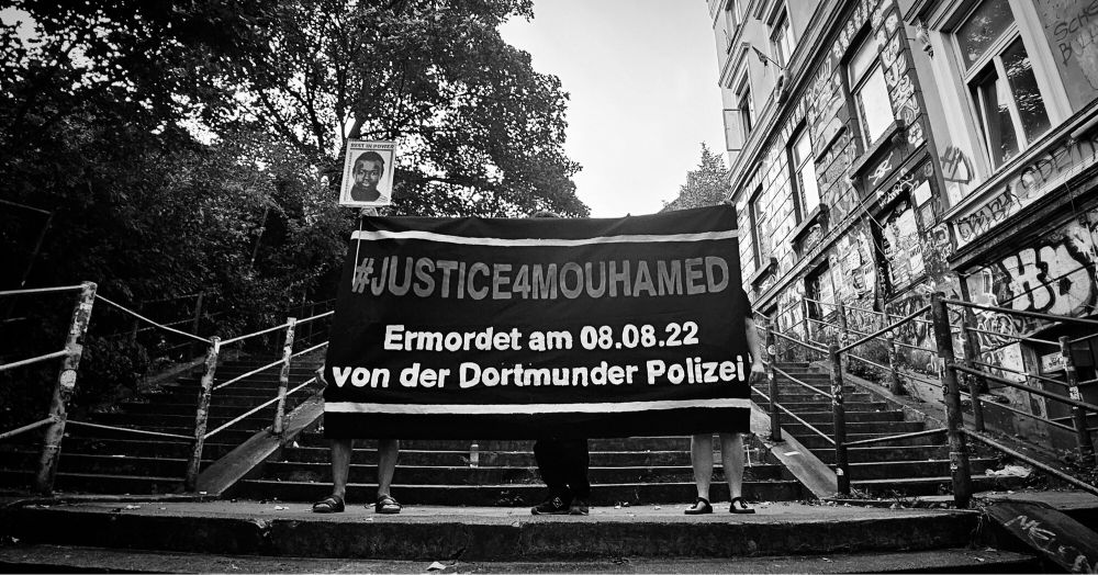 Das Foto zeigt die Balduintreppe, auf der drei Personen stehen. Diese halten ein Banner im die Höhe, auf welchem zu lesen ist: #Justice4Mouhamed Ermordet am 08.08.22 von der Dortmunder Polizei.