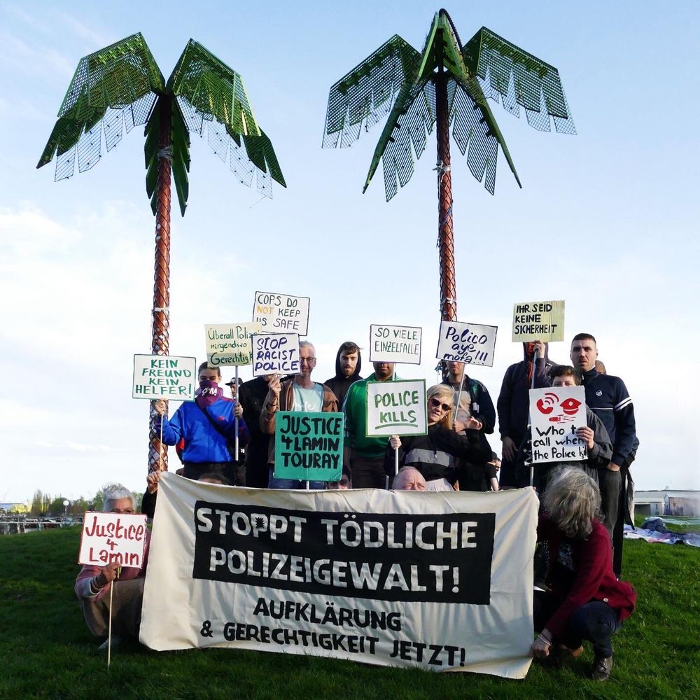 Solidaritätsfoto in Gedenken an Lamin Touray. Zu sehen ist eine kleine Gruppe von Menschen, die im Park Fiction ein Banner & Schilder in die Höhe halten auf denen u.a. zu lesen ist "Justice 4 Lamin Touray". Auf dem Banner steht: "Stoppt tödliche Polizeigewalt! Aufklärung & Gerechtigkeit jetzt."