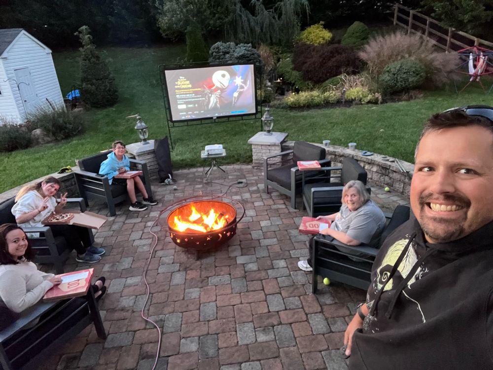 Photograph of a backyard patio with a projector setup and a fire pit going. On the projector is The Nightmare Before Christmas. My family is sitting around the fire pit and I’m in the foreground with a Nightmare Before Christmas hoodie