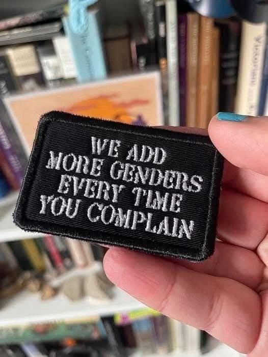 A hand holding a patch. The nail of the thumb is visibly painted turquoise. The patch is made from black fabric and has embroidered white text on it: "WE ADD MORE GENDERS EVERY TIME YOU COMPLAIN"
In the blurry background are bookshelves.