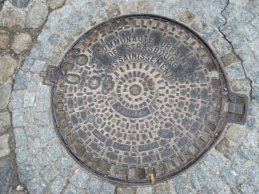 A round manhole cover set in cobblestones that says 
Communate Urbaine de Strasbourg 
Assignissement