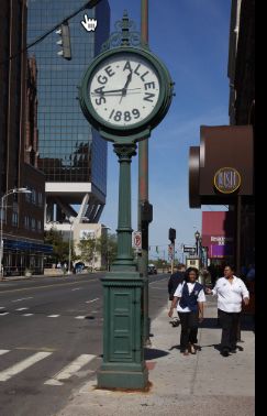Main street HArtford Connecticut, large green stylized clock stands on the side of the road, wording of Sage-Allen and 1889 display on the face, peopel walk by 