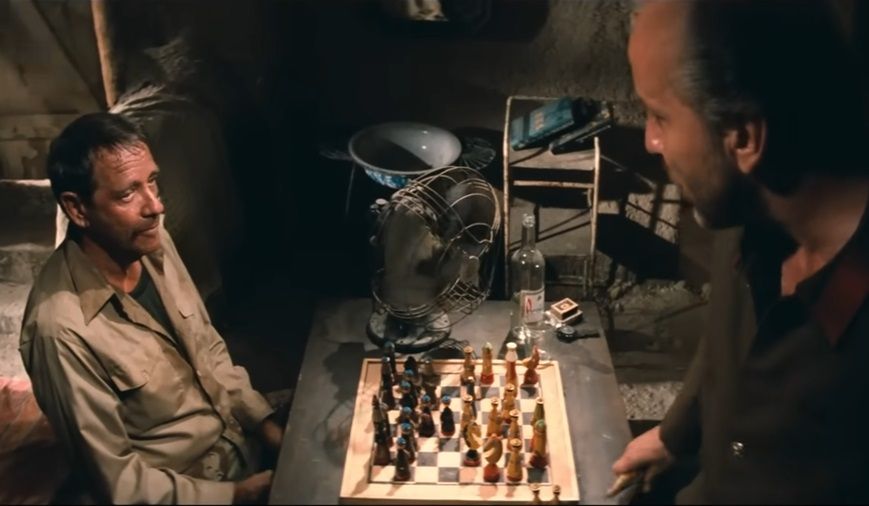 In a scene from Rambo III, Richard Crenna, looking dirty and haggard in a khaki shirt, sits at a table where a chess game is in progress and looks up at Mark de Jonge, who looms over the table in the extreme foreground.