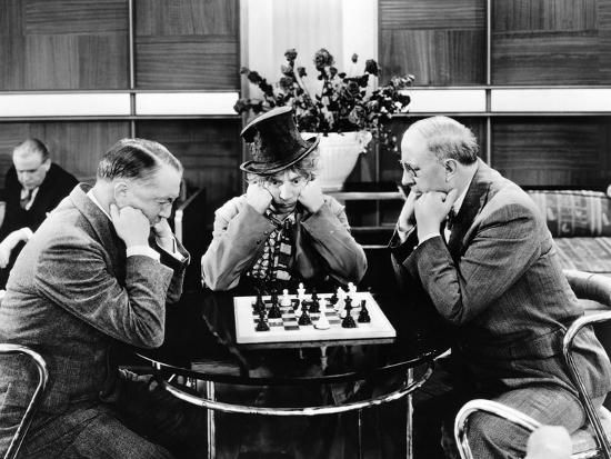 In a scene from Monkey Business, two middle-aged men in suits sit at a table playing chess, each with his elbows on the table and resting his chin on his hands. Harpo Marx sits at the table watching the game from the side, mimicking the players' posture.